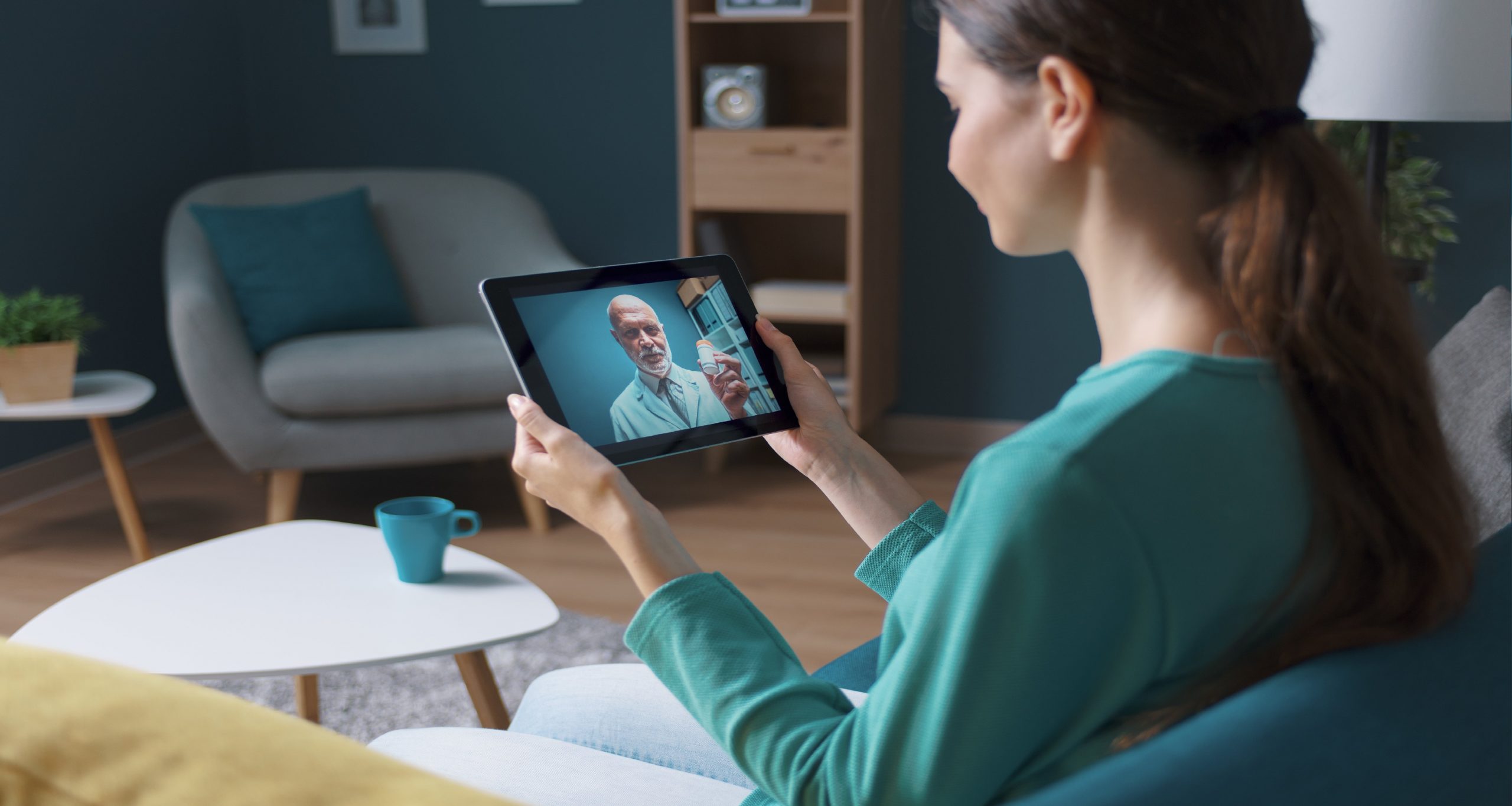 A woman sitting on a couch, holding a tablet, engaged in a conversation with a doctor. Example of telemedicine.