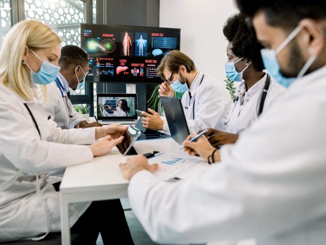 Group of multiethnical healthcare workers in masks, with digital tablets and laptop, meeting in hospital boardroom.
