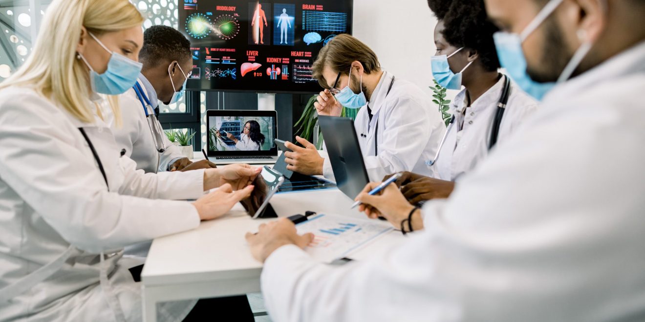 Group of multiethnical healthcare workers in masks, with digital tablets and laptop, meeting in hospital boardroom.