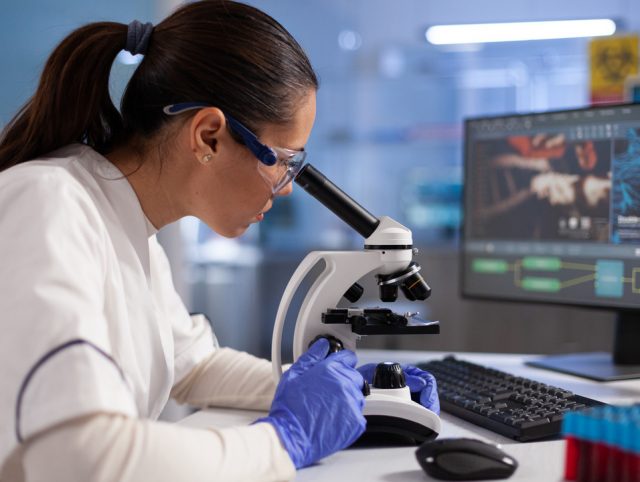 A woman in lab coat conducting microscope analysis. Visual for clinical trials management.