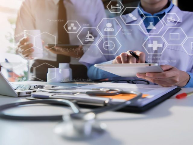 A doctor using a tablet. Laptop, documentation and stethoscope in front of him, an array of medical icons displayed on the screen.