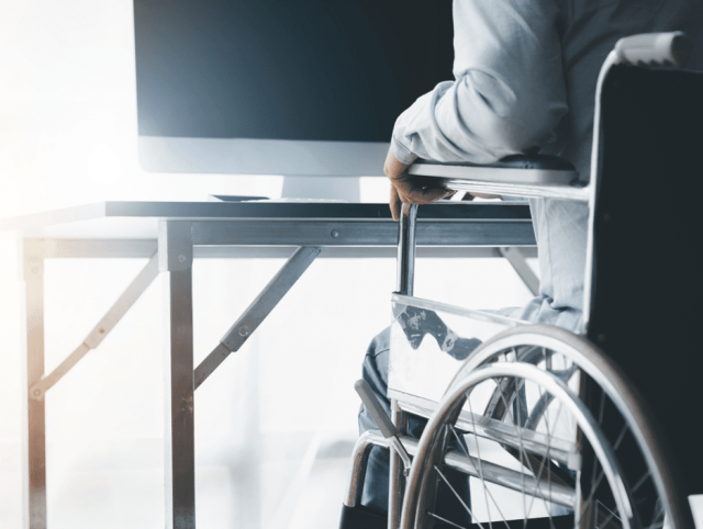 A person in a wheelchair is seated at a desk, using a computer; visual for screen readers & voice recognition software.