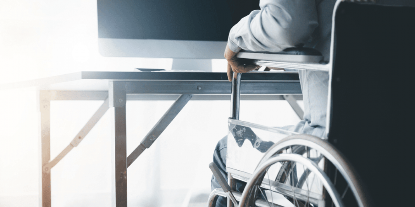 A person in a wheelchair is seated at a desk, using a computer; visual for screen readers & voice recognition software.