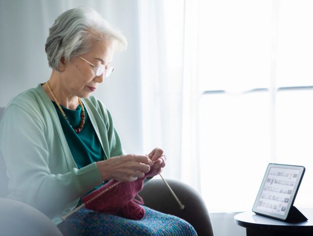 An elderly woman sits in a chair knitting, with a tablet on the table, illustrating the integration of smart home technology.