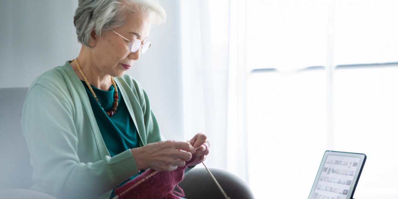 An elderly woman sits in a chair knitting, with a tablet on the table, illustrating the integration of smart home technology.