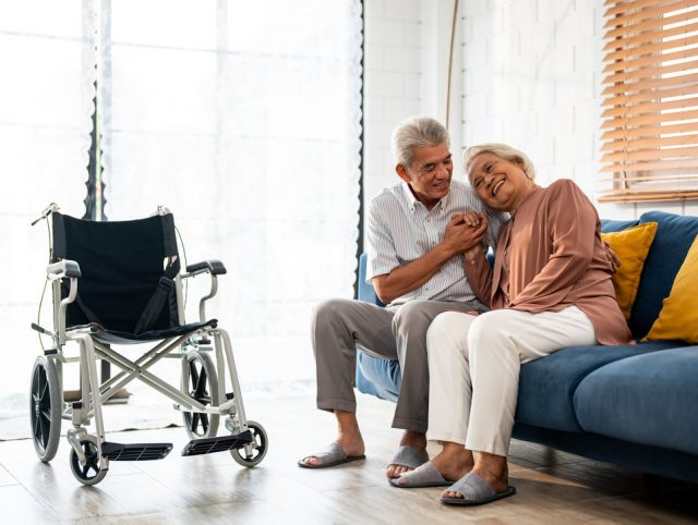 An elderly couple seated on a couch with a wheelchair in the background.