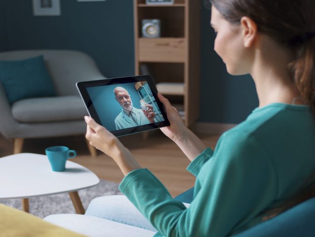 A woman sitting on a couch, holding a tablet, engaged in a conversation with a doctor. Example of telemedicine.