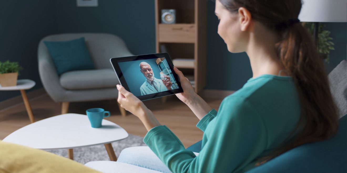 A woman sitting on a couch, holding a tablet, engaged in a conversation with a doctor. Example of telemedicine.